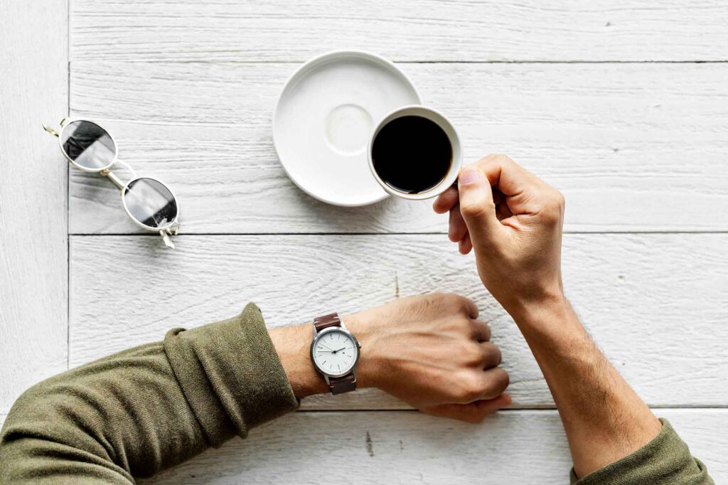 Mãos sob uma mesa. Uma das mãos está segurando uma xícara de café preto. Evitar cafeína após o entardecer costuma nos ajudar a dormir bem à noite.