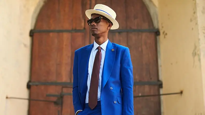 Homem utilizando um chapéu do tipo velejador/boater. Ele também utiliza um traje formal composto por calça e terno nas cores azul, camisa social, gravata e óculos de sol.