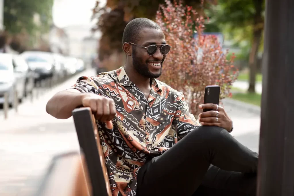 Homem sentado em um banco de praça, sorrindo, olhando seu smartphone