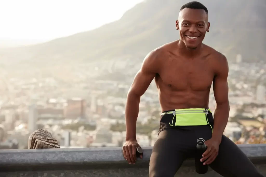 Homem de pele negra, sem camisa, ao ar livre. Ele está sentado, utilizando um short típico de quem pratica corrida.