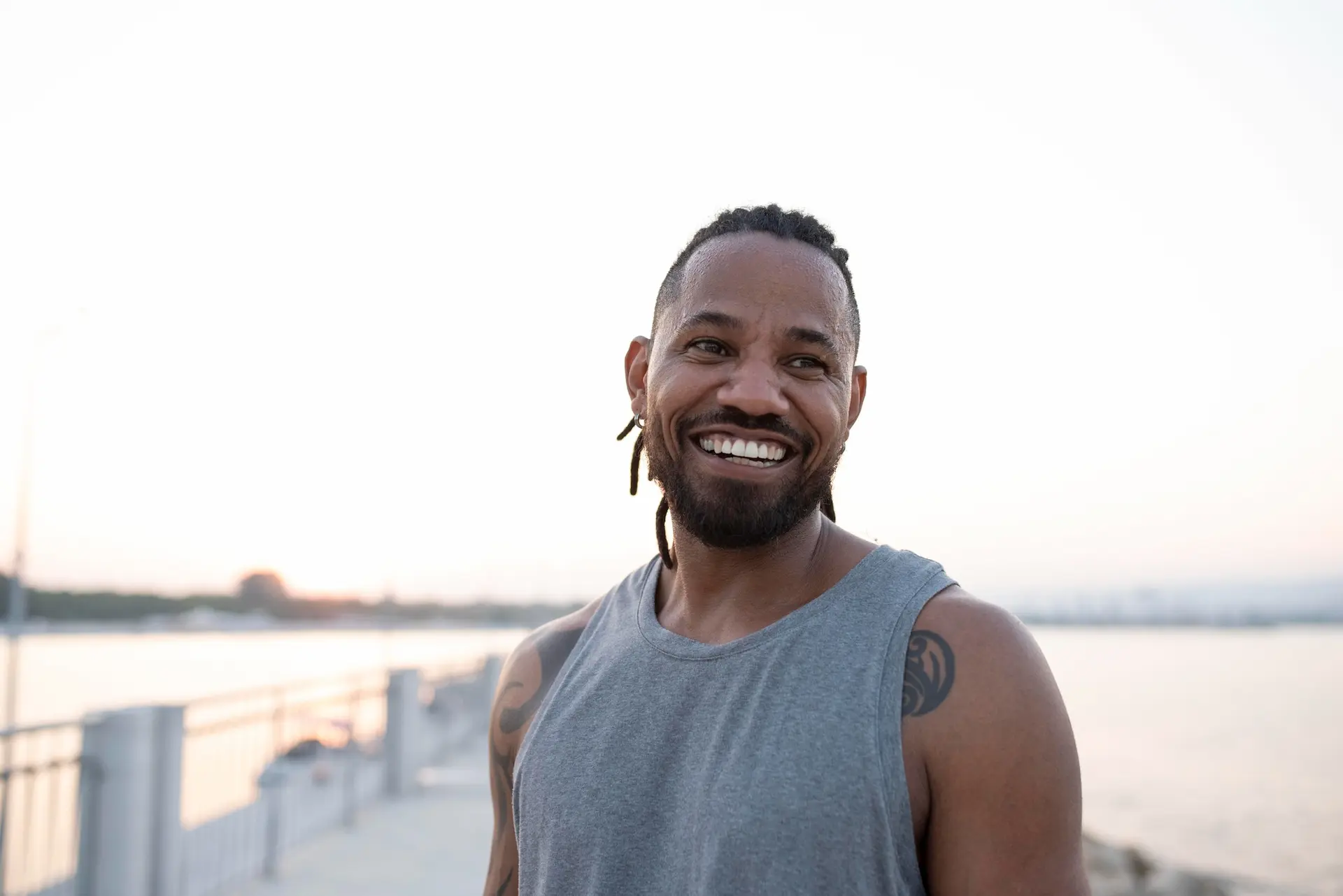 Homem de pele negra sorrindo. Ele está em um local ao ar livre utilizando uma roupa que indica que o mesmo estava se exercitando. Atividades físicas ao ar livre é um ótimo exemplo de hábitos saudáveis.