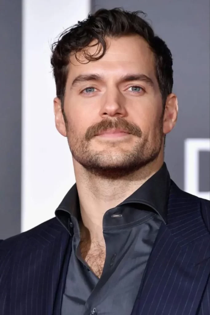 Ator Henry Cavill com barba por fazer, mas com um bigode mais destacado e robusto.