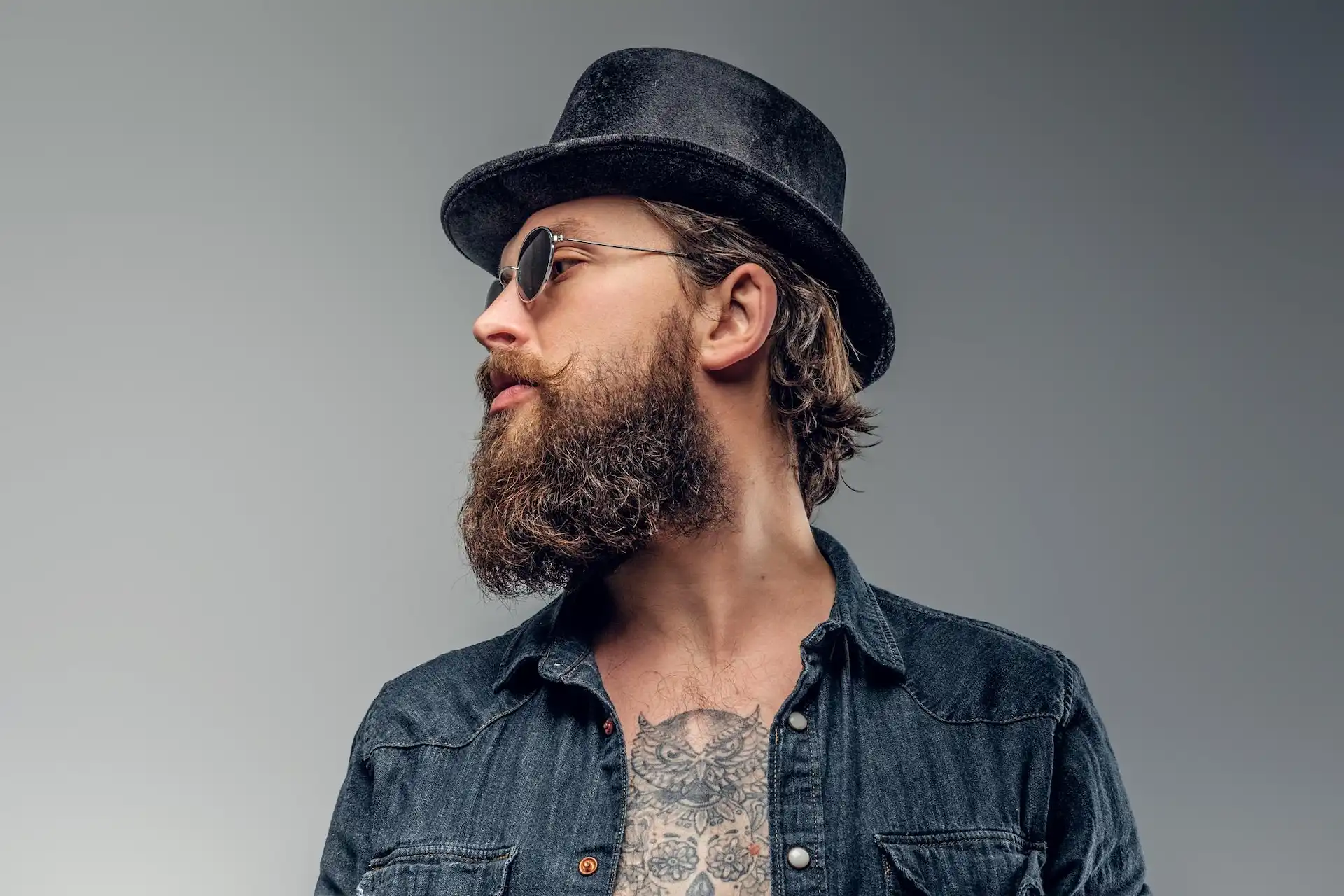 Homem com barba cheia, usando óculos de sol, chapéu preto e jaqueta jeans.