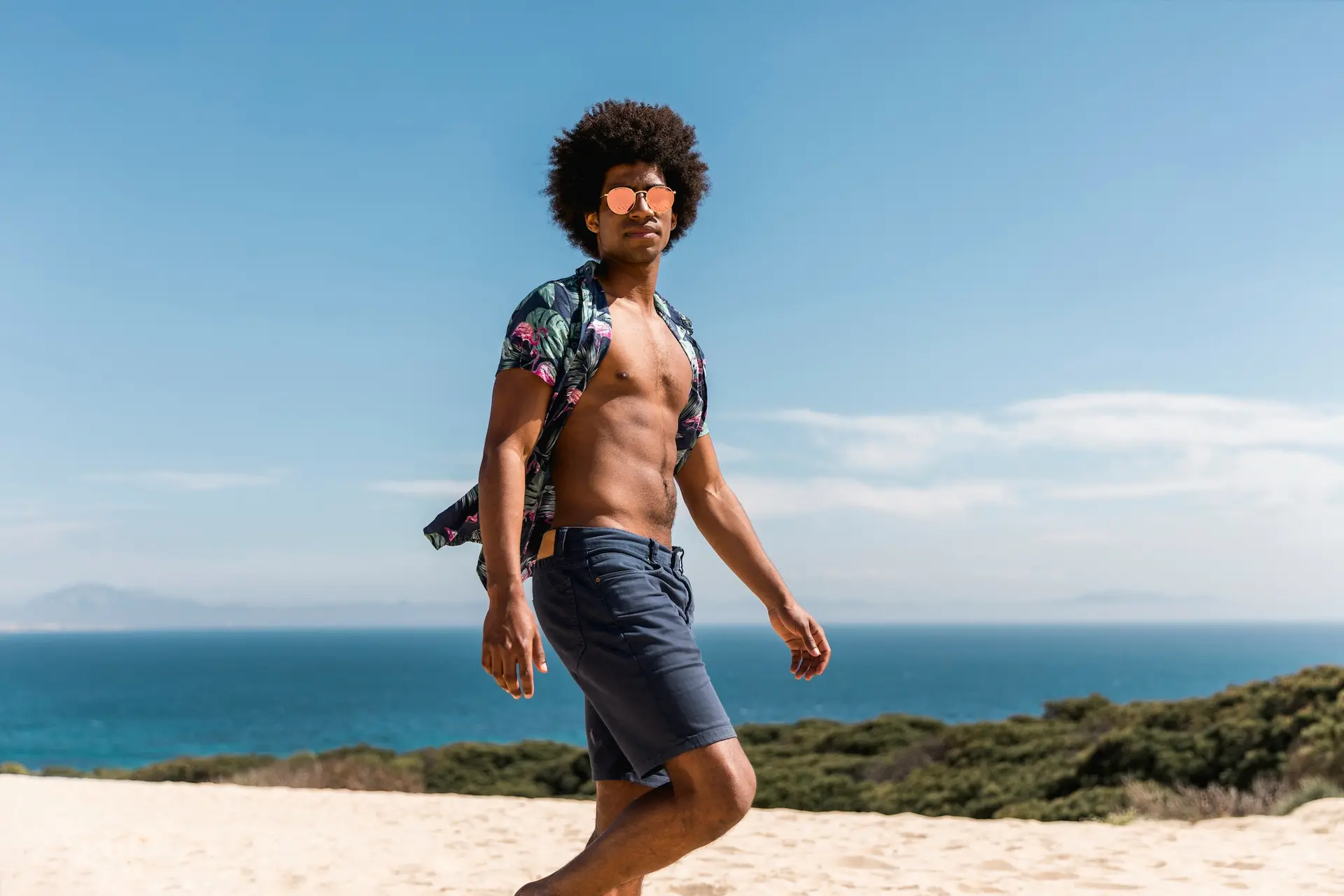 Homem utilizando um look masculino com bermuda e camisa estampada na praia.