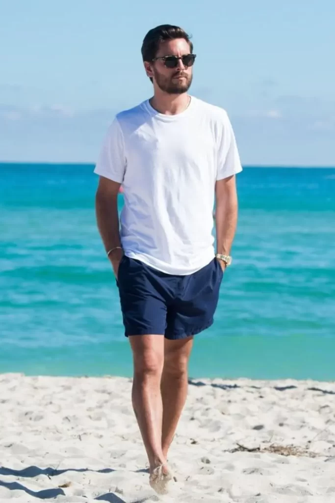 Homem utilizando um look masculino básico na praia, com camiseta, bermuda e óculos de sol.