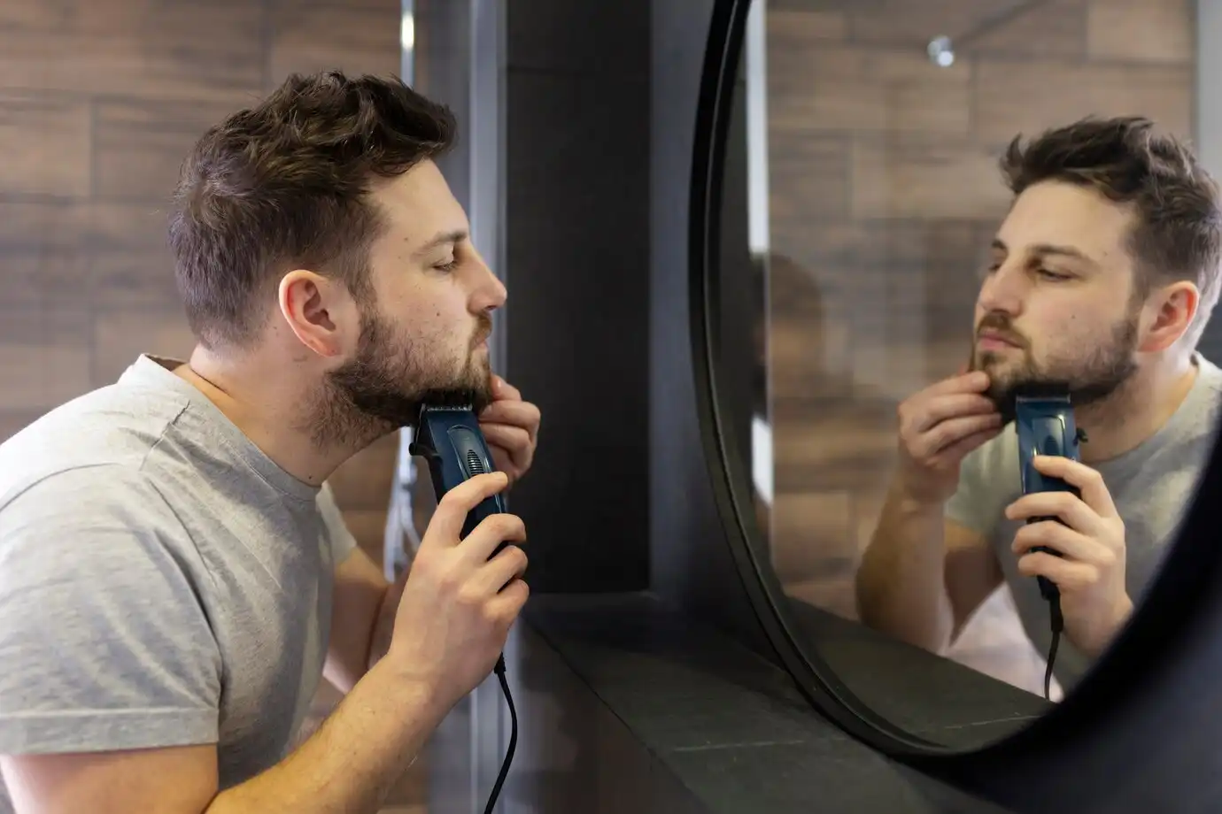 Homem diante de um espelho, aparando a barba com um aparador elétrico.