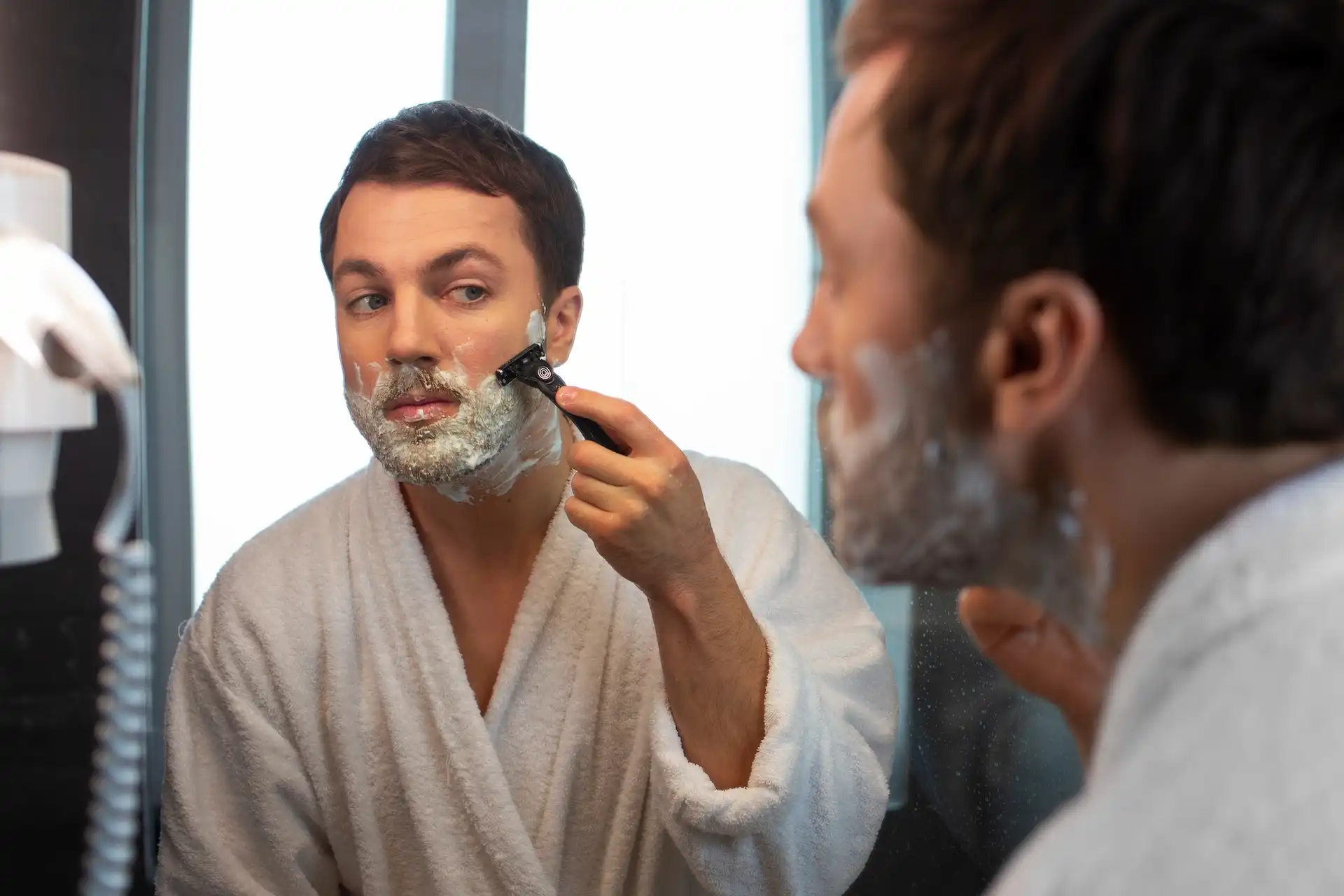 Homem fazendo a barba usando espuma de barbear em frente ao espelho.