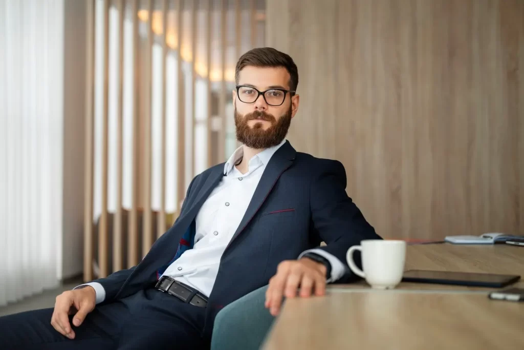 Homem sentado em uma cadeira em um ambiente de trabalho. Ele usa um traje elegante composto por calça e camisa social, e blazer. Em seu rosto, há um óculos de grau.