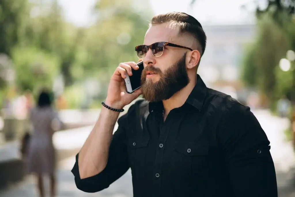 Homem com a barba longa impecável falando ao celular na rua. Hidratar a barba é fundamental para a aparência e saúde dos fios e pele.