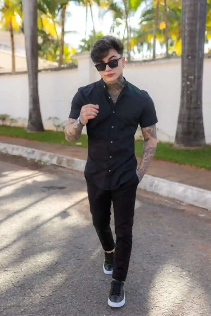 Homem vestido todo de preto. Ele usa tênis casual, calça jeans e camisa. Além disso, ele está usando óculos de sol.