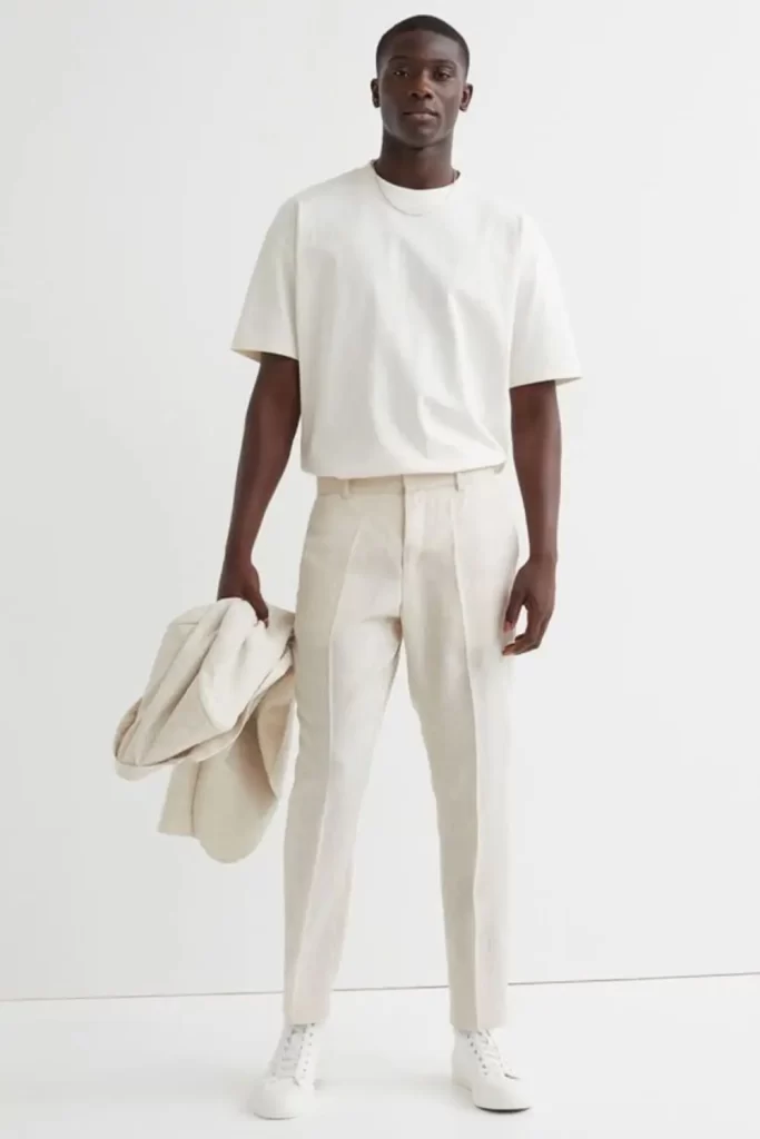 Homem usando um look monocromático masculino branco. Ele usa tênis casual branco, calça de tecido leve e camiseta. Nas mãos, ele segura um casaco.