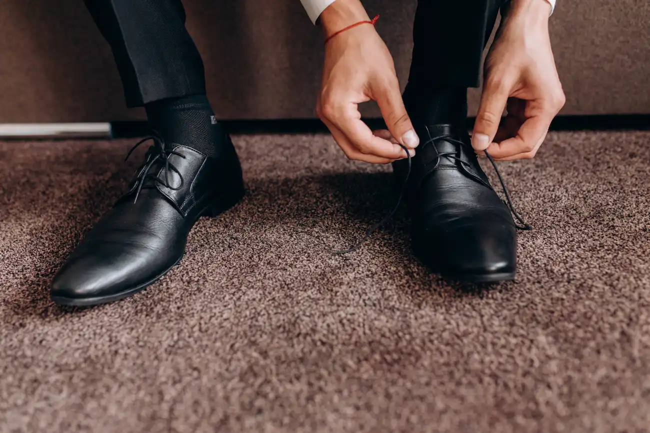 A imagem mostra de perto, os pés de um homem calçando sapatos pretos. As mãos dele estão amarrando um dos sapatos.