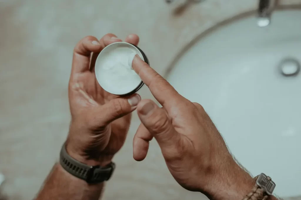 A imagem mostra duas mãos masculinas manuseando um balm para barba. Uma das mãos está com o dedo indicador com uma pequena quantidade do produto.