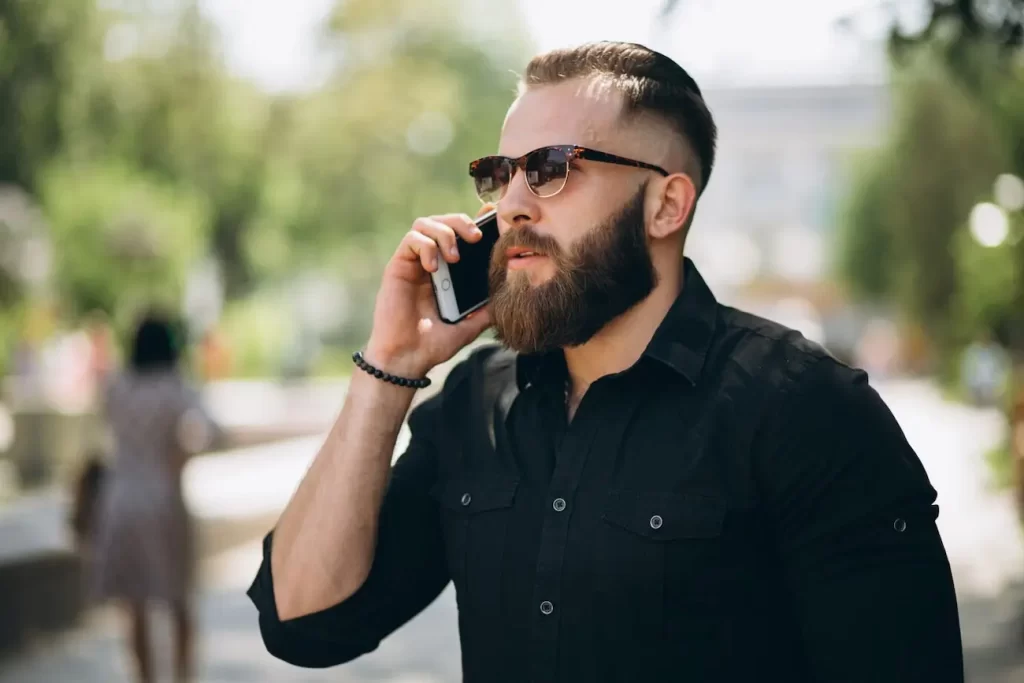 Homem com barba cheia falando no celular em uma rua.