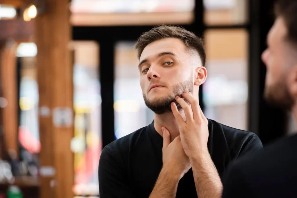 Homem observando sua barba com falhas no espelho.