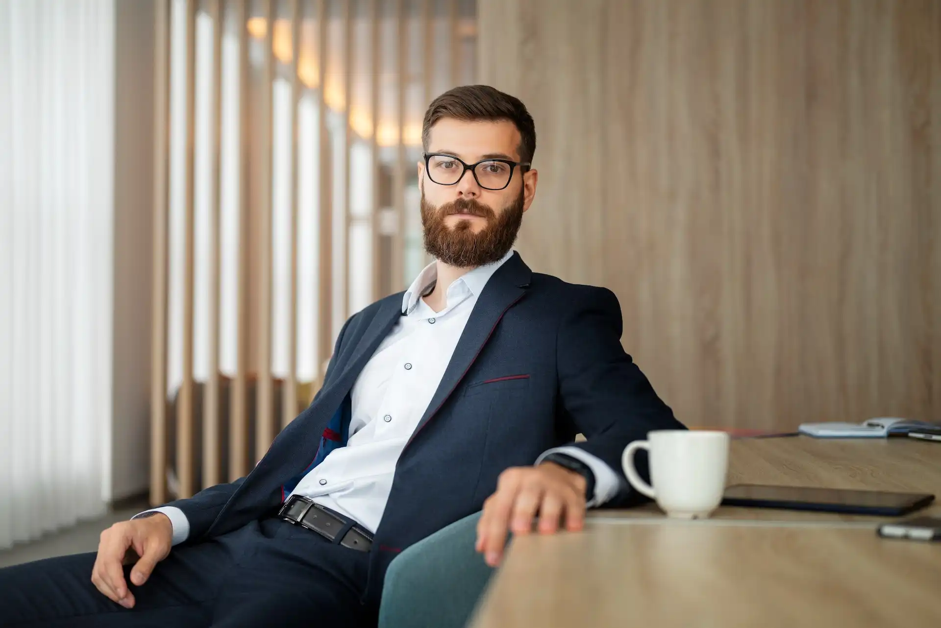 Homem elegante sentado ao lado de uma mesa. Ele está usando uma roupa formal composta por: calça e camisa social, e blazer. Em seu rosto, há um óculos de grau.