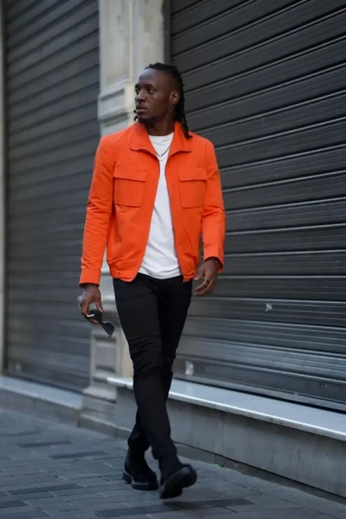 Homem usando camiseta branca, jaqueta laranja, calça e bota preta.