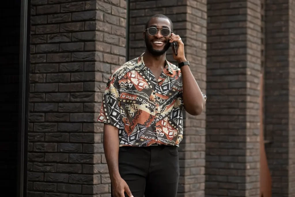 Homem usando calça de alfaiataria preta, camisa larga com estampas em tons marrons e óculos de sol. Ele está em uma rua falando ao celular.