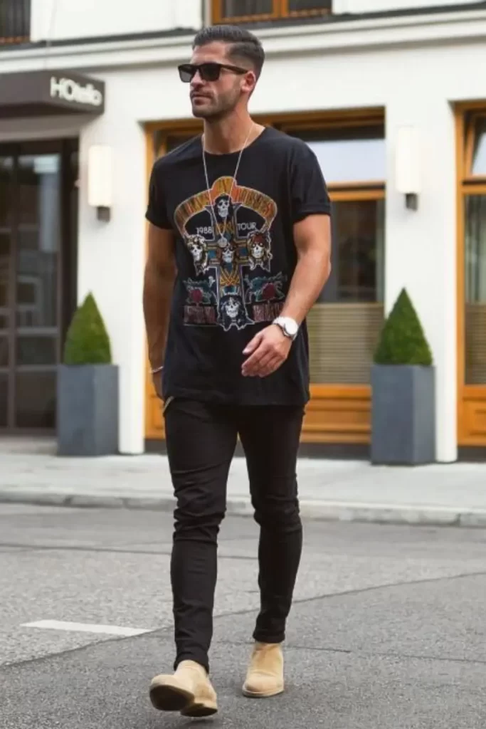 Homem vestido conforme um estilo rock masculino básico. Ele usa calça jeans, camiseta da banda Guns N’ Roses e acessórios (pulseira, relógio, colar e óculos de sol).