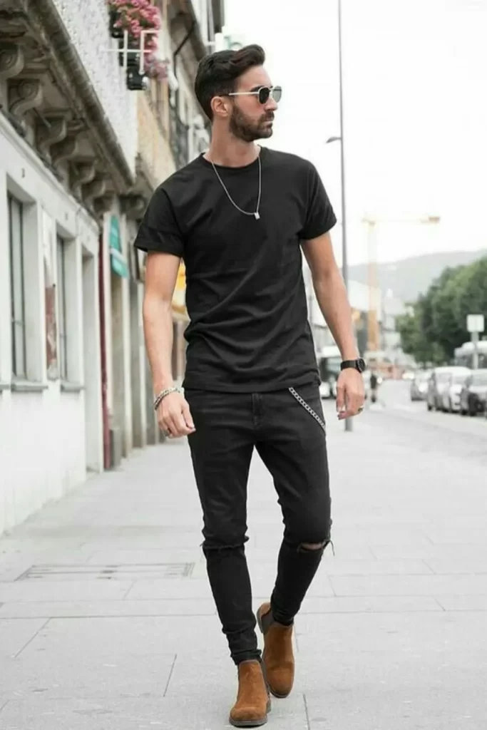 Homem vestido conforme um estilo rock masculino básico, com calça, camiseta minimalista e botas de couro. Além disso, ele usa pulseira, relógio, colar e óculos de sol.