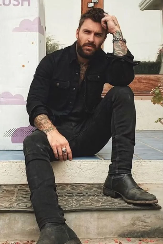 Homem usando calça jeans, camiseta, jaqueta jeans e botas. Todas as peças na cor preta. Ele também usa uma pulseira de corrente e um anel.