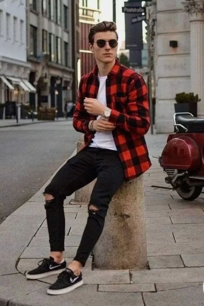 Homem usando um estilo rock grunge, com tênis, calça rasgada, camiseta branca e camisa xadrez vermelha sobre a camiseta. Ele também usa óculos de sol, uma pulseira de couro e relógio.