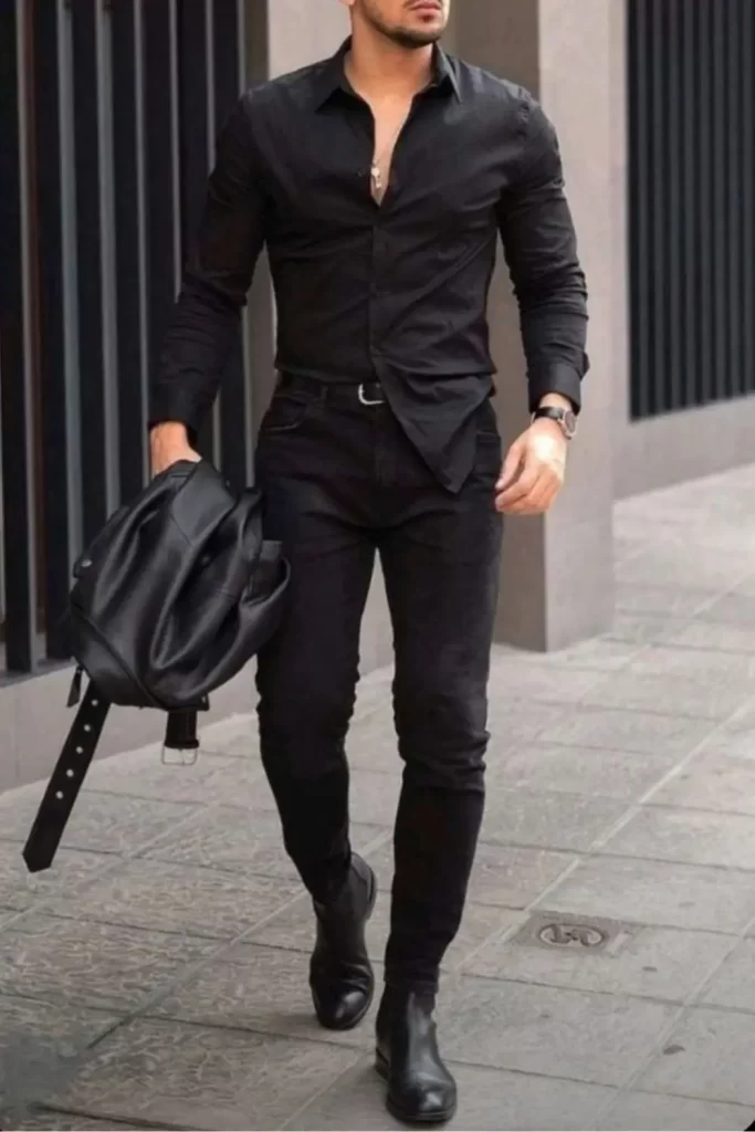 Homem usando um estilo rock masculino mais elegante e formal. Ele usa botas de couro, calça jeans e camisa social. Todas as peças na cor preta. Ele está segurando uma jaqueta de couro em uma das mãos, e usa um relógio elegante .