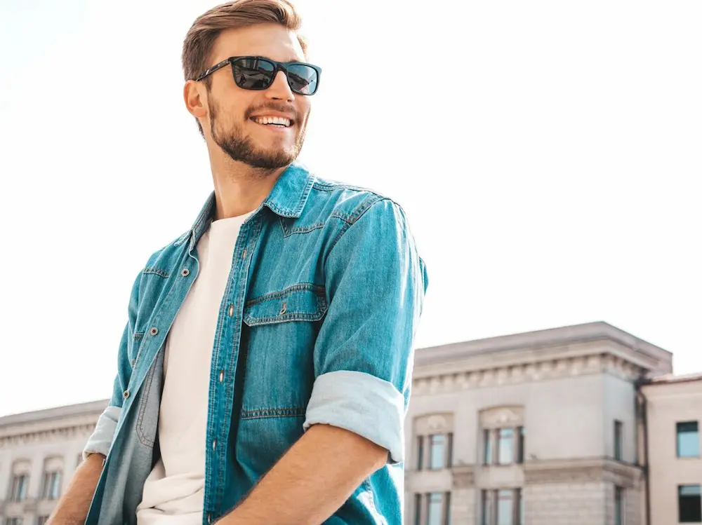 Homem usando camiseta branca, jaqueta jeans azul e óculos de sol.