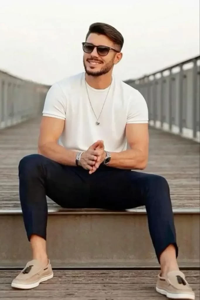 Homem usando camiseta branca, calça chino azul-escuro, mocassim e óculos de sol. Nos pulsos, relógio e pulseira.