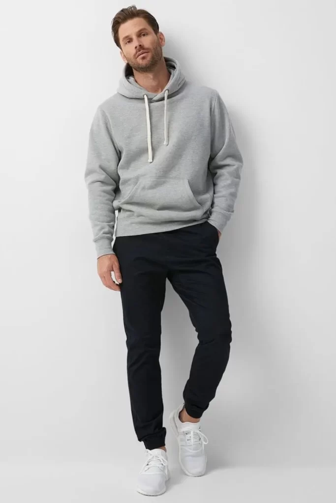 Homem usando um look esportivo. Ele usa tênis branco, calça jogger preta e moletom cinza.