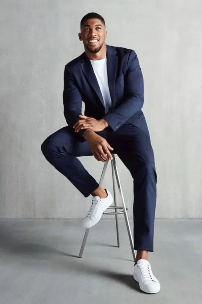 Homem usando calça de alfaiataria azul-marinho, camiseta branca, blazer na mesmo cor da calça e tênis casual branco.